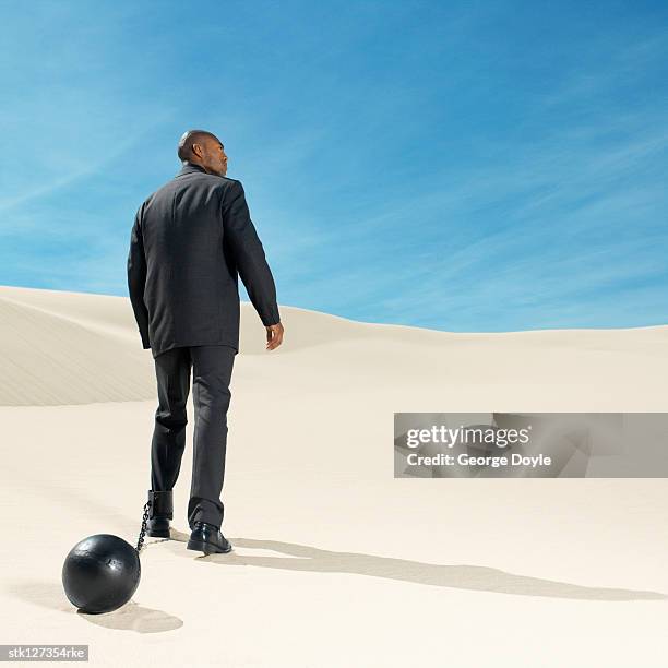 businessman wearing ball and chain in desert rear view, low angle view - restraining device stock pictures, royalty-free photos & images