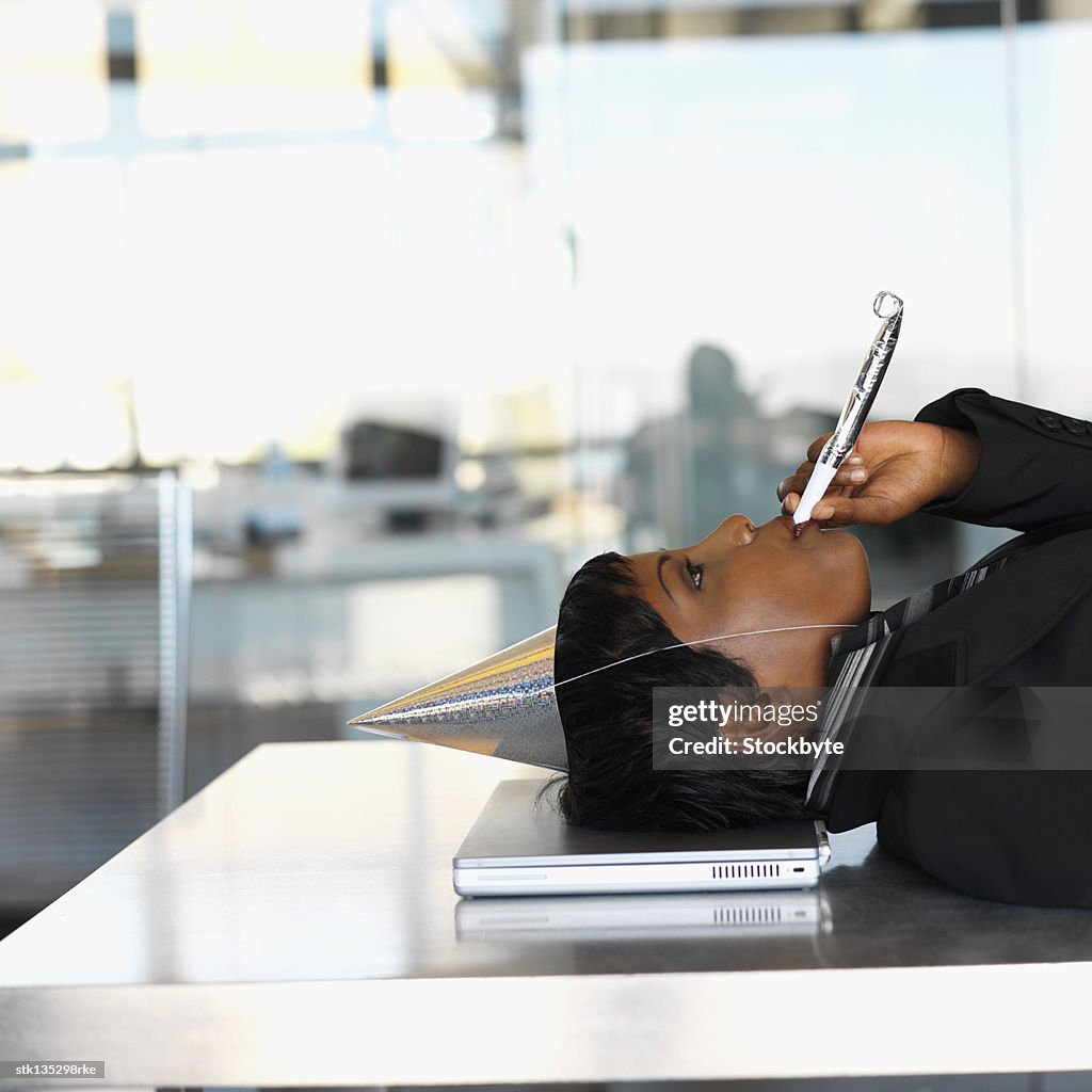 Side view of a businesswoman wearing a party hat and blowing a whistle with her head resting on laptop