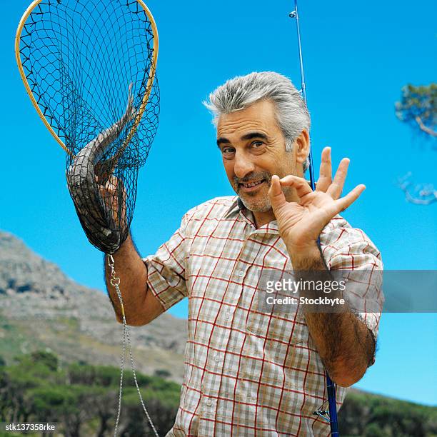 front view portrait of a mature man holding a fishing rod and a fish in a net giving a signal of happiness - signal stock-fotos und bilder