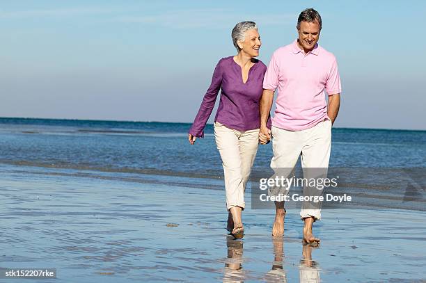 elderly couple holding hands walking on the beach smiling - purple shirt stock pictures, royalty-free photos & images