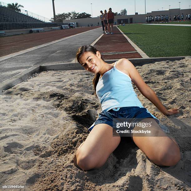female long jump athlete (18-21) kneeling in sandpit smiling after jump - womens field event stockfoto's en -beelden