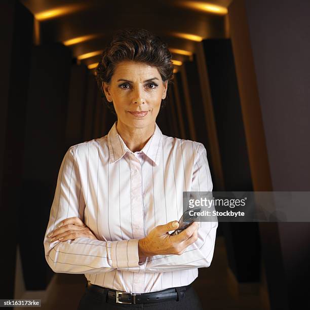 portrait of a woman standing with her arms folded in a hallway holding mobile phone in her hand - folded blouse stock pictures, royalty-free photos & images