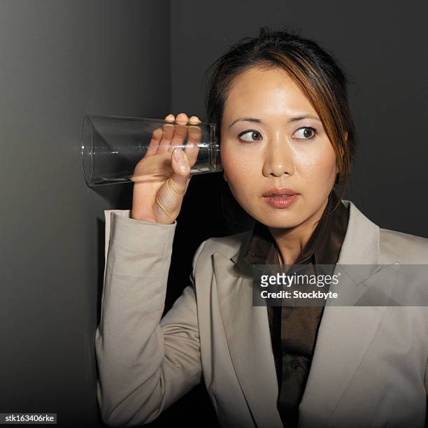 businesswoman eaves-dropping in office by holding glass to the wall portrait close-up - eaves - fotografias e filmes do acervo