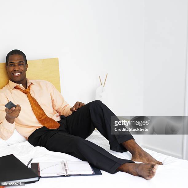 businessman sitting on a bed relaxing and using television remote control - control photos et images de collection