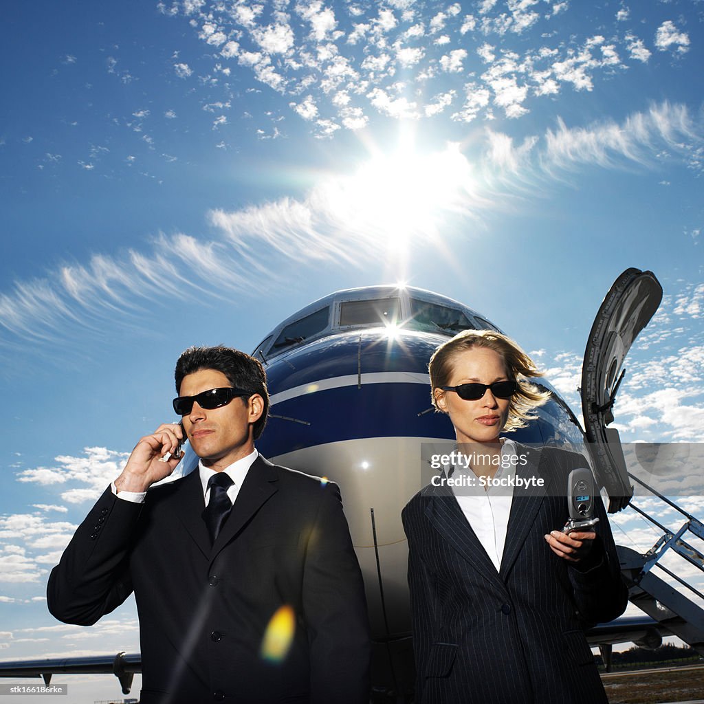 Business executives standing in front of an airplane and talking on mobile phones