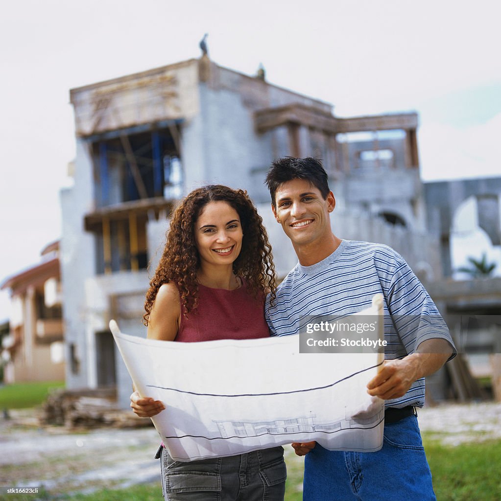 Portrait of a couple looking at blueprint and house under construction in background