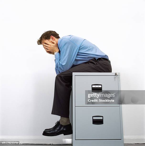 low angle view of man sitting on top a file cabinet clutching his face - file cabinet stock-fotos und bilder