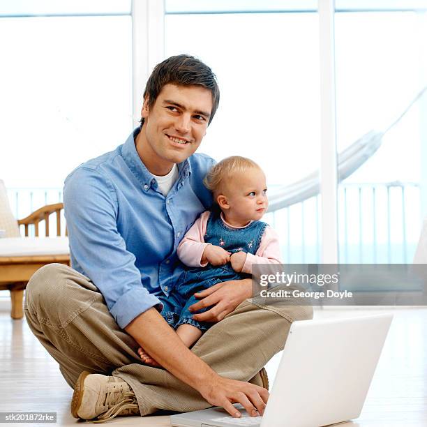 father sitting holding his baby daughter (12-18 months) while working on his computer - genderblend stock-fotos und bilder