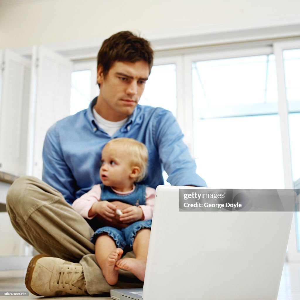 Father sitting holding his baby daughter (12-18 months) while working on his computer