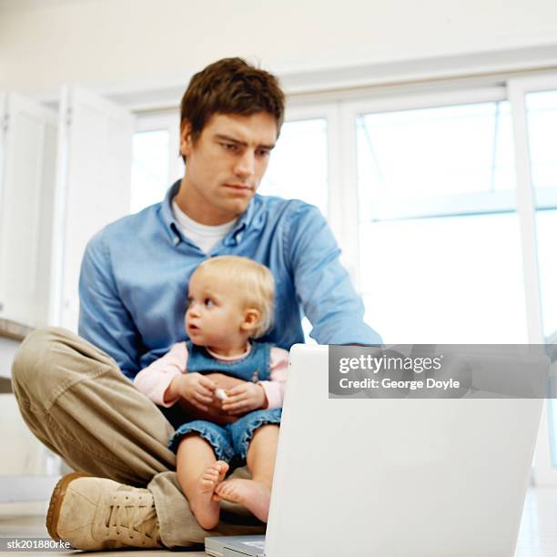 father sitting holding his baby daughter (12-18 months) while working on his computer - genderblend stock-fotos und bilder