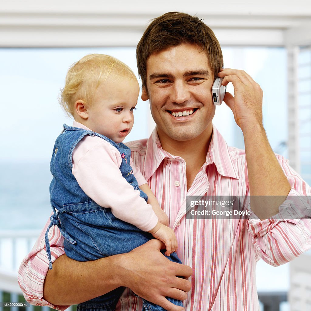 Father holding baby daughter (12-18 months) while talking on mobile phone