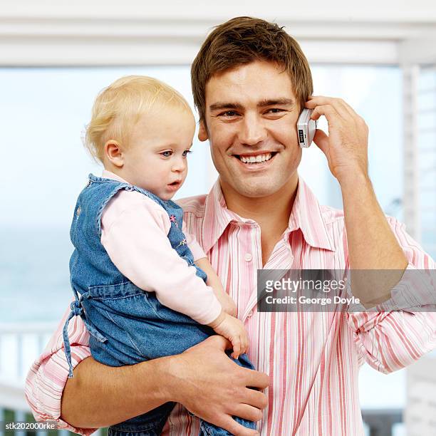 father holding baby daughter (12-18 months) while talking on mobile phone - genderblend stock pictures, royalty-free photos & images