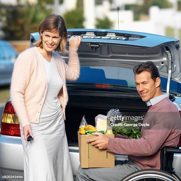 man sitting in wheelchair and wife bringing in groceries from car - closing car boot fotografías e imágenes de stock