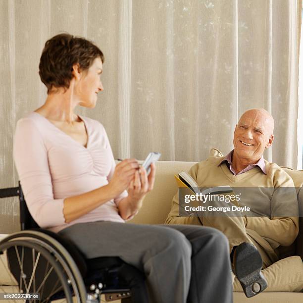 woman sitting in a wheelchair holding a personal digital assistant and man sitting on sofa reading a book - silver surfer stock pictures, royalty-free photos & images