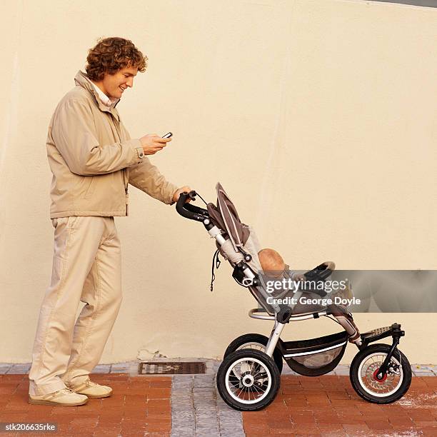 mother pushing her baby (12-18 months) in a buggy while talking on her mobile phone - genderblend stock-fotos und bilder