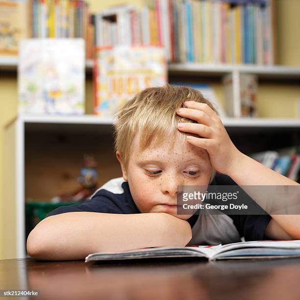 boy (7-10) with down syndrome reading a book in a classroom - books on shelf stockfoto's en -beelden