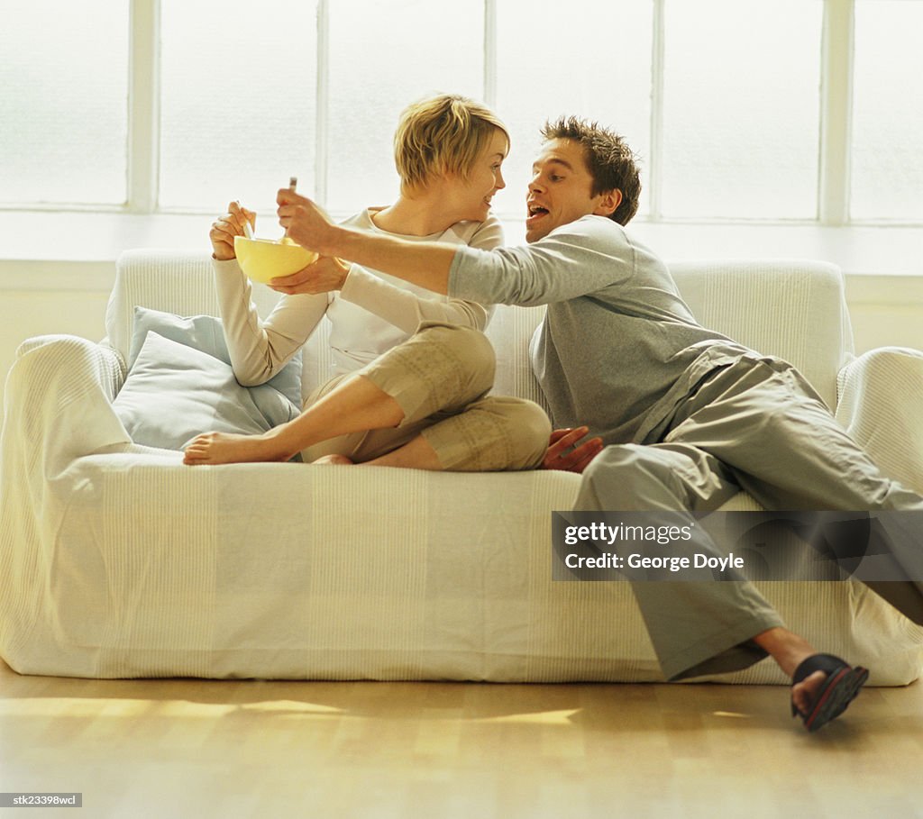 A playful couple sitting on a sofa eating from a bowl