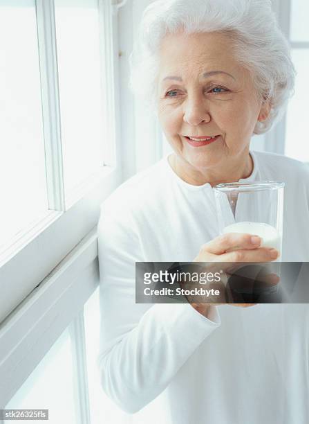 portrait of an elderly woman standing next to a window with a glass of milk - next imagens e fotografias de stock