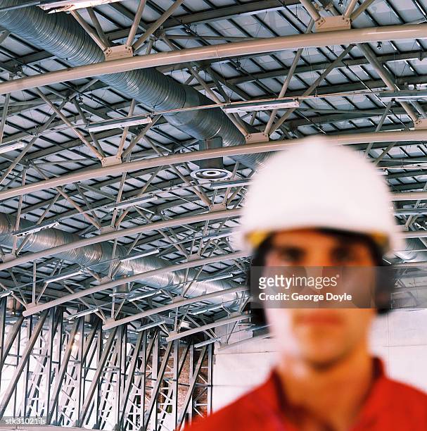 close-up of a young man wearing a hard hat standing under metal scaffolding (blurred) - hat stock-fotos und bilder