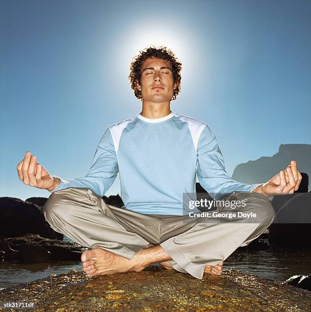 low angle view of a young man sitting cross legged in a meditative pose on a rock at the beach - brian may signs copies of we will rock you at virigin megastore september 28 2004 stockfoto's en -beelden