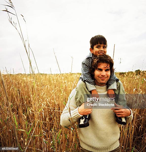 portrait of a young man walking through a field with his son riding piggyback - genderblend stock-fotos und bilder