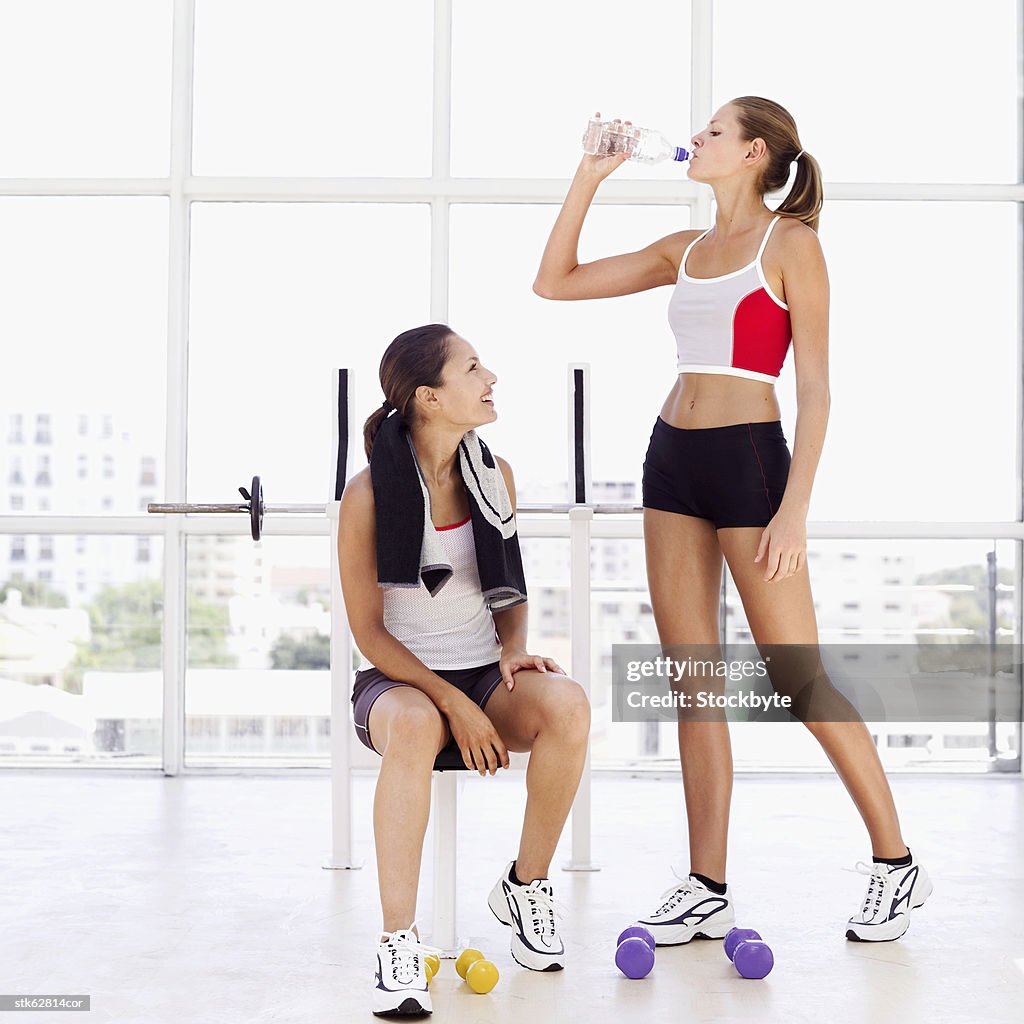 Woman standing and drinking water from a bottle with another woman sitting on a bench