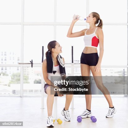 Woman standing and drinking water from a bottle with another woman sitting on a bench Woman standing and drinking water from a bottle with another woman sitting on a bench