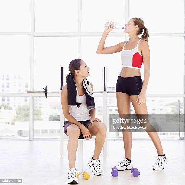 woman standing and drinking water from a bottle with another woman sitting on a bench - aother stock pictures, royalty-free photos & images