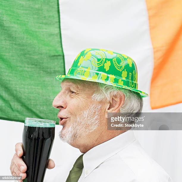 side view of an elderly man wearing a plastic hat drinking beer beside the irish flag - hat stock-fotos und bilder