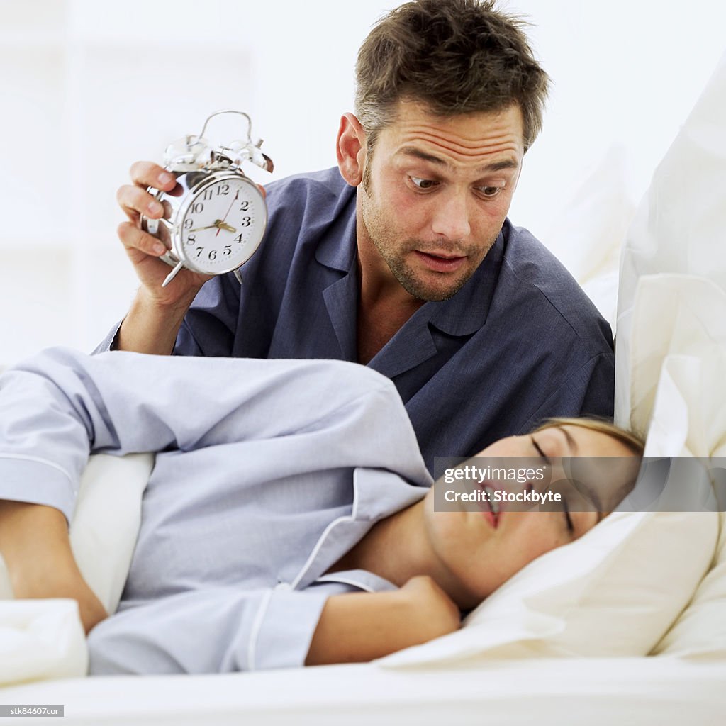 Shocked man holding an alarm clock and looking at his wife