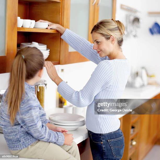 girl (8-9) sitting on the kitchen counter top and watching her mother put away dishes - her stock pictures, royalty-free photos & images