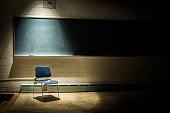 An Empty School Chair in a Dark, Shadowy Classroom - in Front of a Chalkboard with a Single Beam of Light Overhead