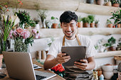 Asian male florist, owner of small business flower shop, using digital tablet while working on laptop against flowers and plants. Checking stocks, taking customer orders, selling products online. Daily routine of running a small business with technology