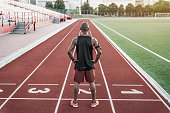 Athlete standing at the start line with hands on waist. Rear view of runner standing on running track.