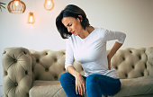 Axial pain. Close-up photo of a hurting woman, who is sitting on a couch and holding her lower back with her left hand.