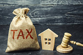 A bag with money and the word tax next to a wooden house. Taxes on real estate, payment. Penalty, arrears. Register of taxpayers for property. Law-abiding, evasion of payment. Court law