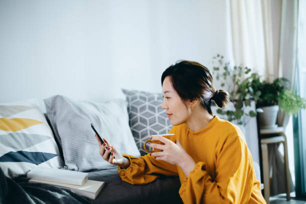 beautiful smiling young asian woman chilling at cozy home, sitting on the floor by the sofa, enjoying a cup of coffee and using smartphone - women stock pictures, royalty-free photos & images