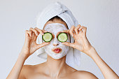 Beautiful young woman with facial mask on her face holding slices of fresh cucumber