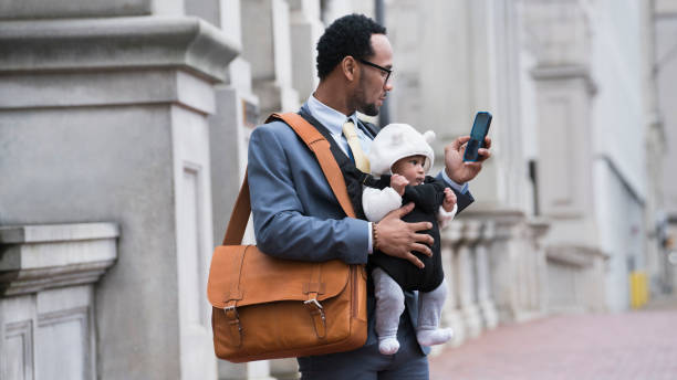 black businessman with son in baby carrier texting on cell phone - working black dad stock pictures, royalty-free photos & images