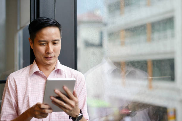 business man using a tablet next to a window - middle aged asian man using a tablet stock pictures, royalty-free photos & images
