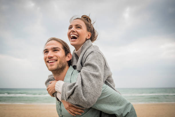caucasian man carrying woman piggyback on beach - couple stock pictures, royalty-free photos & images