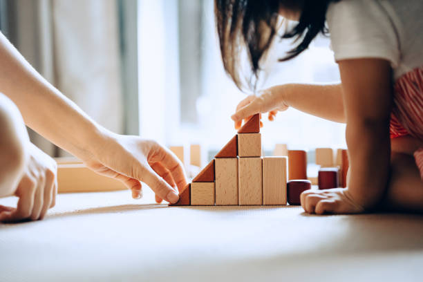 close up of mother and little daughter sitting on the floor playing with wooden building blocks together and enjoying family bonding time at home - parents stock pictures, royalty-free photos & images