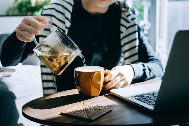 close up of young asian woman pouring a cup of tea from a stylish transparent tea pot into a cup. starting a great day ahead with a cup of hot tea while working at home on laptop in the fresh morning against sunlight. healthy lifestyle concept - tea stock pictures, royalty-free photos & images