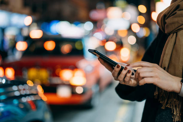 close up of young woman using mobile app device on smartphone to arrange taxi ride in downtown city street, with illuminated busy city traffic scene during rush hour with traffic congestion in the evening - using phone in street stock pictures, royalty-free photos & images