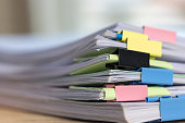 Close up stack of paperwork. Pile of unfinished homework assignment stacked in archive with colorful binder paper clips on table waiting to be managed and inspected. Education and business concept.