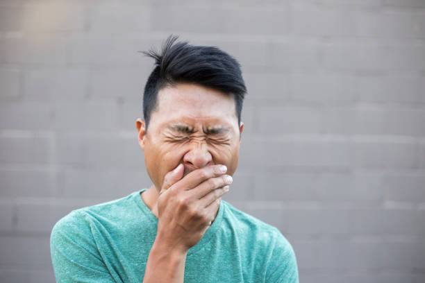 close-up of man with hands covering mouth while yawning against wall - lazy asian man stock pictures, royalty-free photos & images