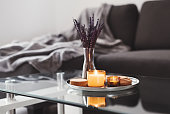 Coffee table design idea: aroma candles and dried lavender bouquet on a metal tray, sofa with grey blanket on background. Simple Scandinavian home decor. Hygge concept
