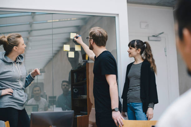 computer programmer writing on adhesive note stuck on glass during meeting with colleagues in office - professional software developer stock pictures, royalty-free photos & images