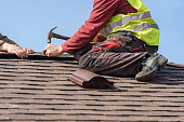 Concept of construction process. Two skilled roofer in special work wear with helmet in hands installing asphalt shingle or roof tile on top of new house