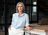 Confident stylish european mature middle aged woman standing at workplace. Stylish older senior businesswoman, 60s gray-haired lady executive leader manager looking at camera in office, portrait.